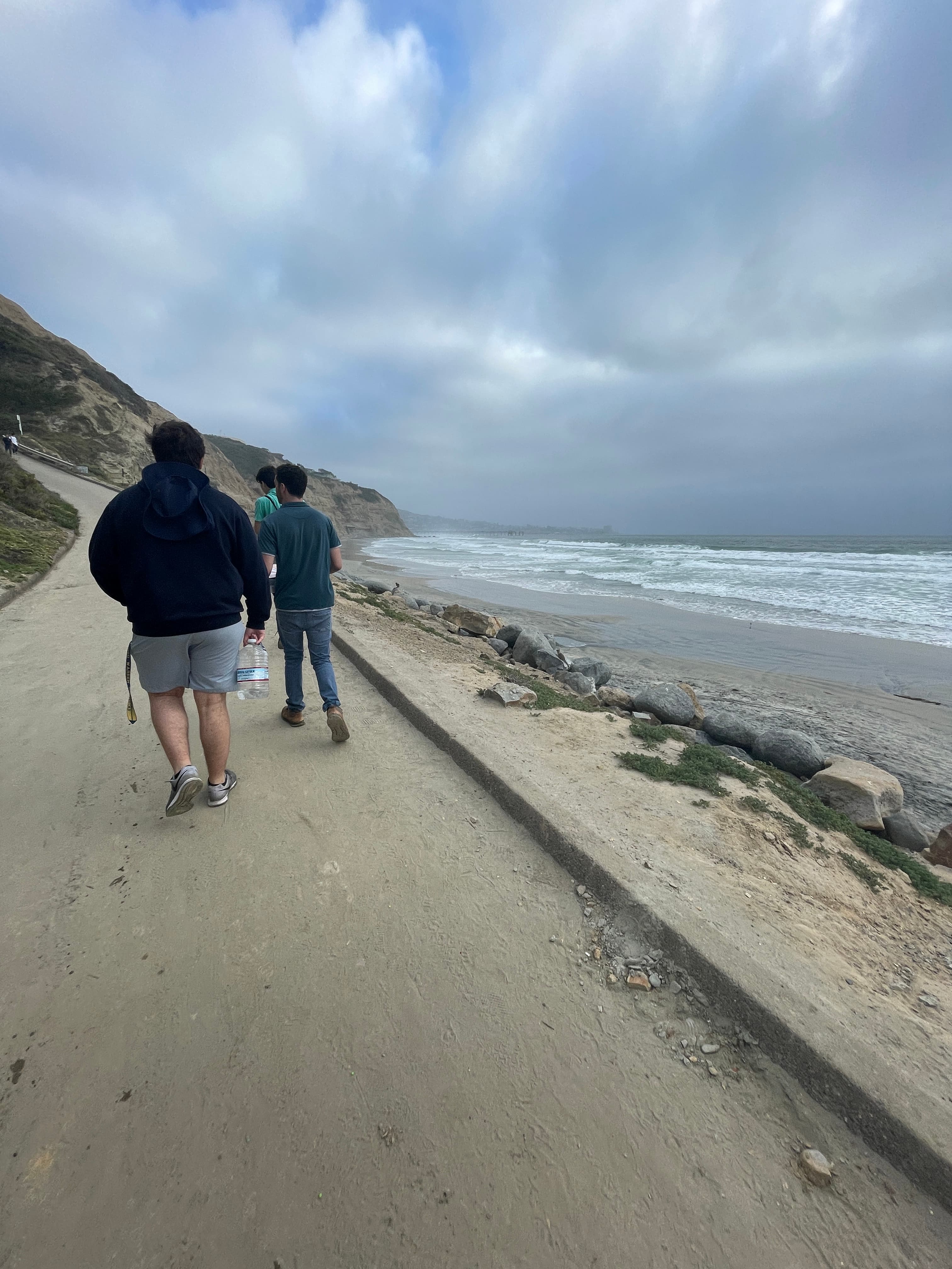 three people, one holding a massive water jug, hiking by the sea